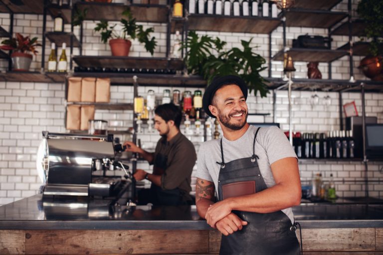 Portrait of cafe owner wearing a hat and apron standing at the counter and looking away. Barista working in background behind the counter making drinks.