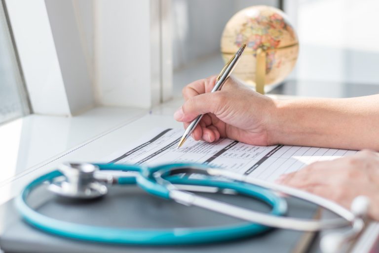 A person filling out a form beside a globe, notebook, and stethoscope