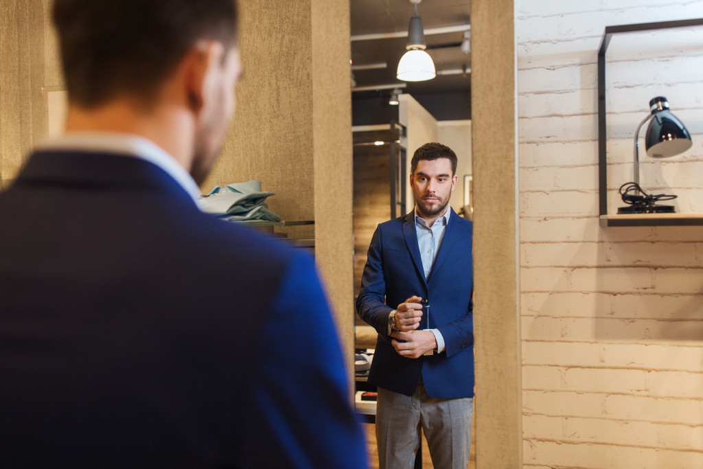 Elegant young man choosing and trying jacket on and looking at the mirror in a clothing store